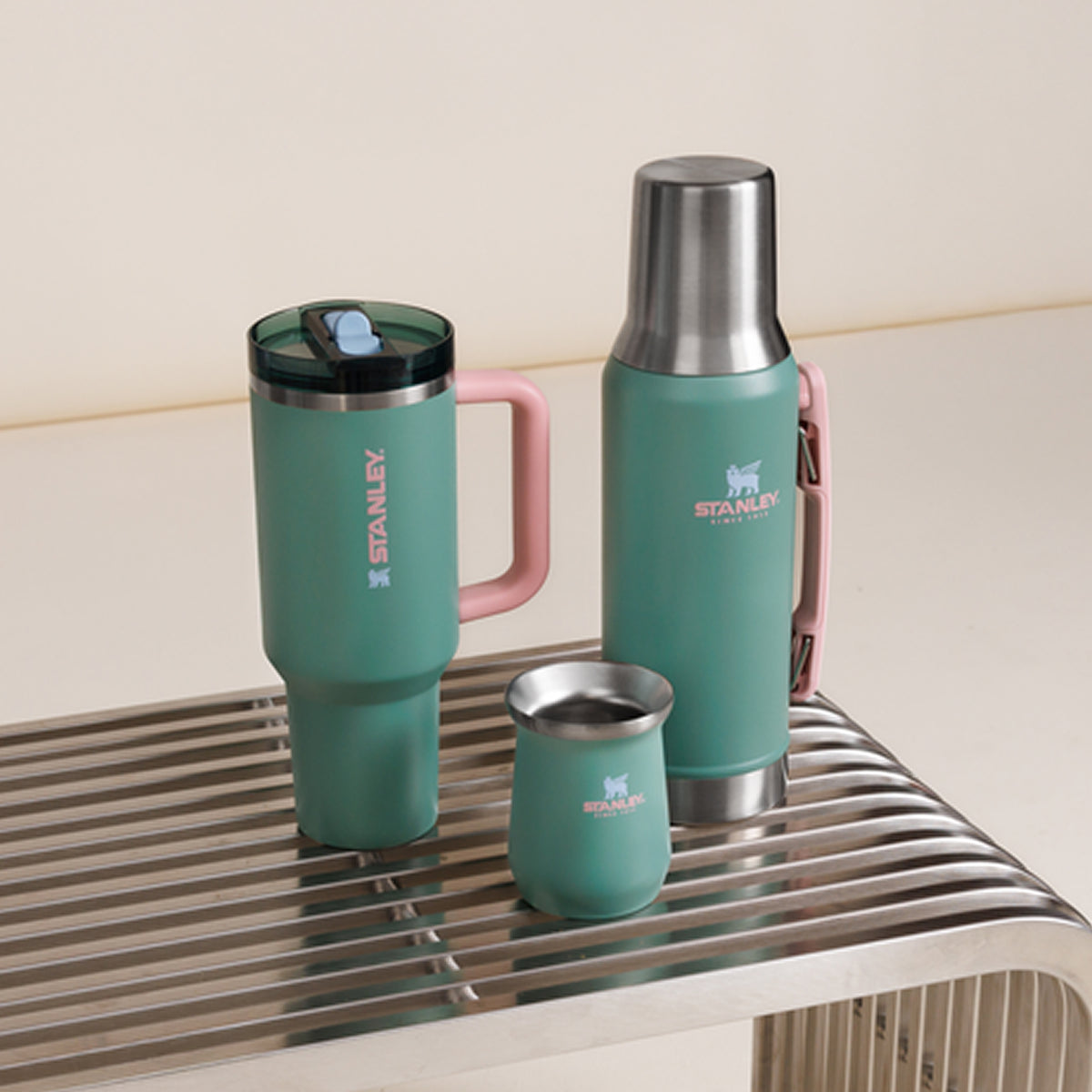 Three Stanley 1913 teal insulated drinkware pieces with pink handles displayed on a metal rack: tumbler, thermos, and small cup.