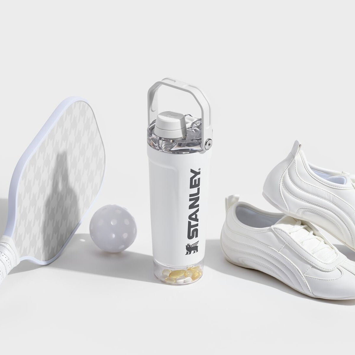 White Stanley 1913 water bottle with pill compartment base, beside a pickleball paddle, ball, and white sneakers.