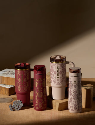 Four Stanley tumblers with lids are arranged on wooden blocks on a tabletop in warm directional light. The tumblers include two deep red designs with gold patterns, one cream and one light blush pink with mauve floral motifs, and one dark brown tumbler with a white handle.