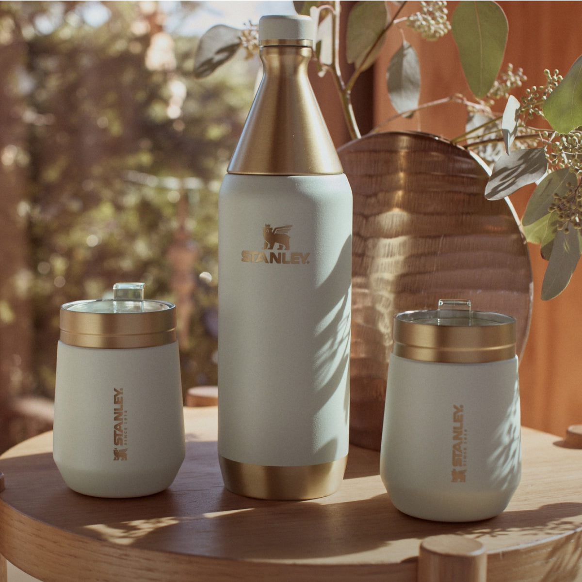 Stanley 1913 cream and gold insulated bottle with two matching tumblers on a wooden table in warm outdoor setting