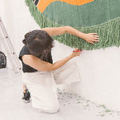 Person painting a large colorful mural on a white wall, crouching near the floor with a brush and paint supplies nearby.
